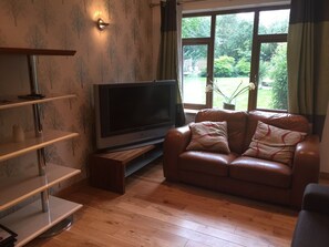 A living room with a flat-screen TV on a wooden stand, a leather sofa with patterned cushions, and a large window with a view of greenery. - The Old Vicarage Lodge (Manchester)