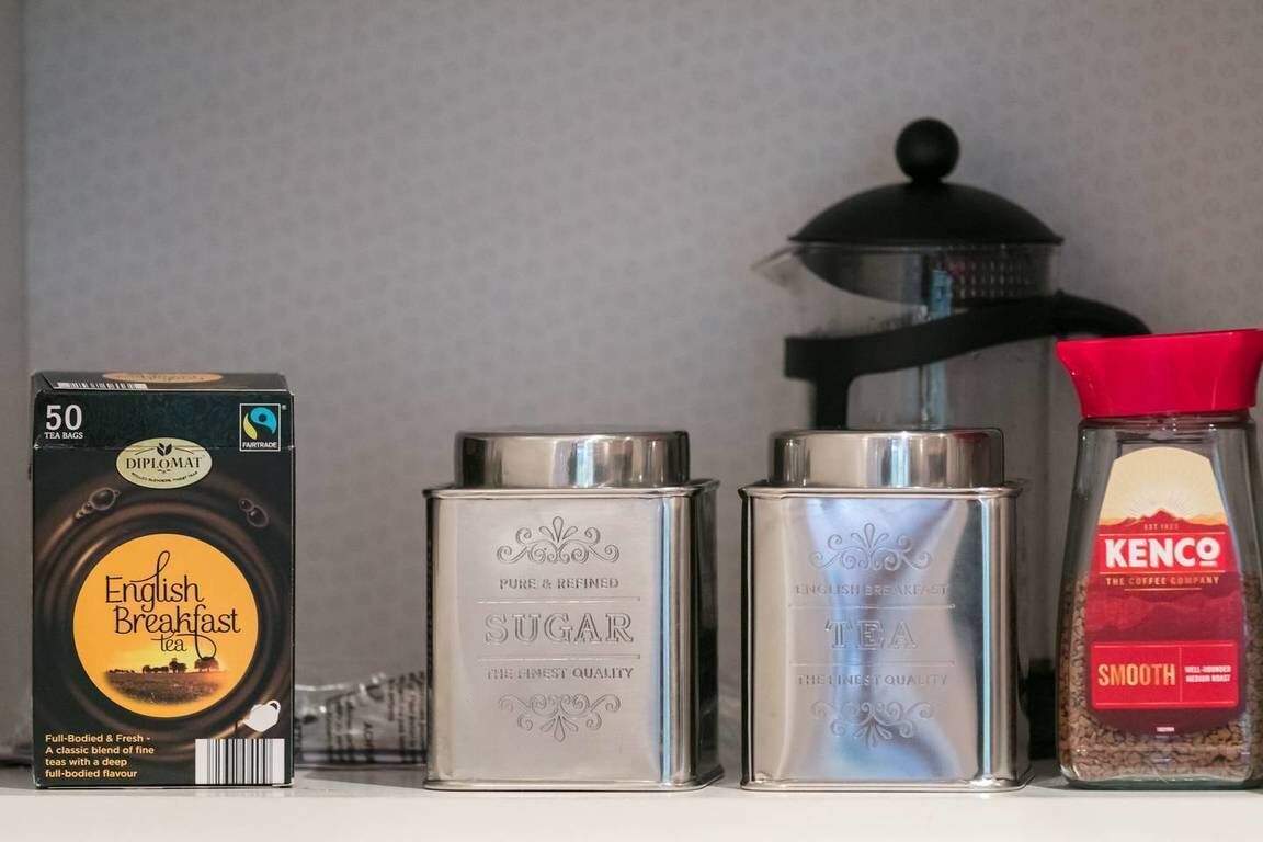 Een plank met een doos English Breakfast thee, twee bakjes met de tekst 'SUGAR' en 'THEE', en een pot Kenco koffie.