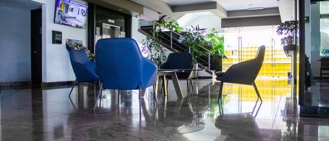 A modern lobby with a reception desk, blue chairs, a staircase, and potted plants.