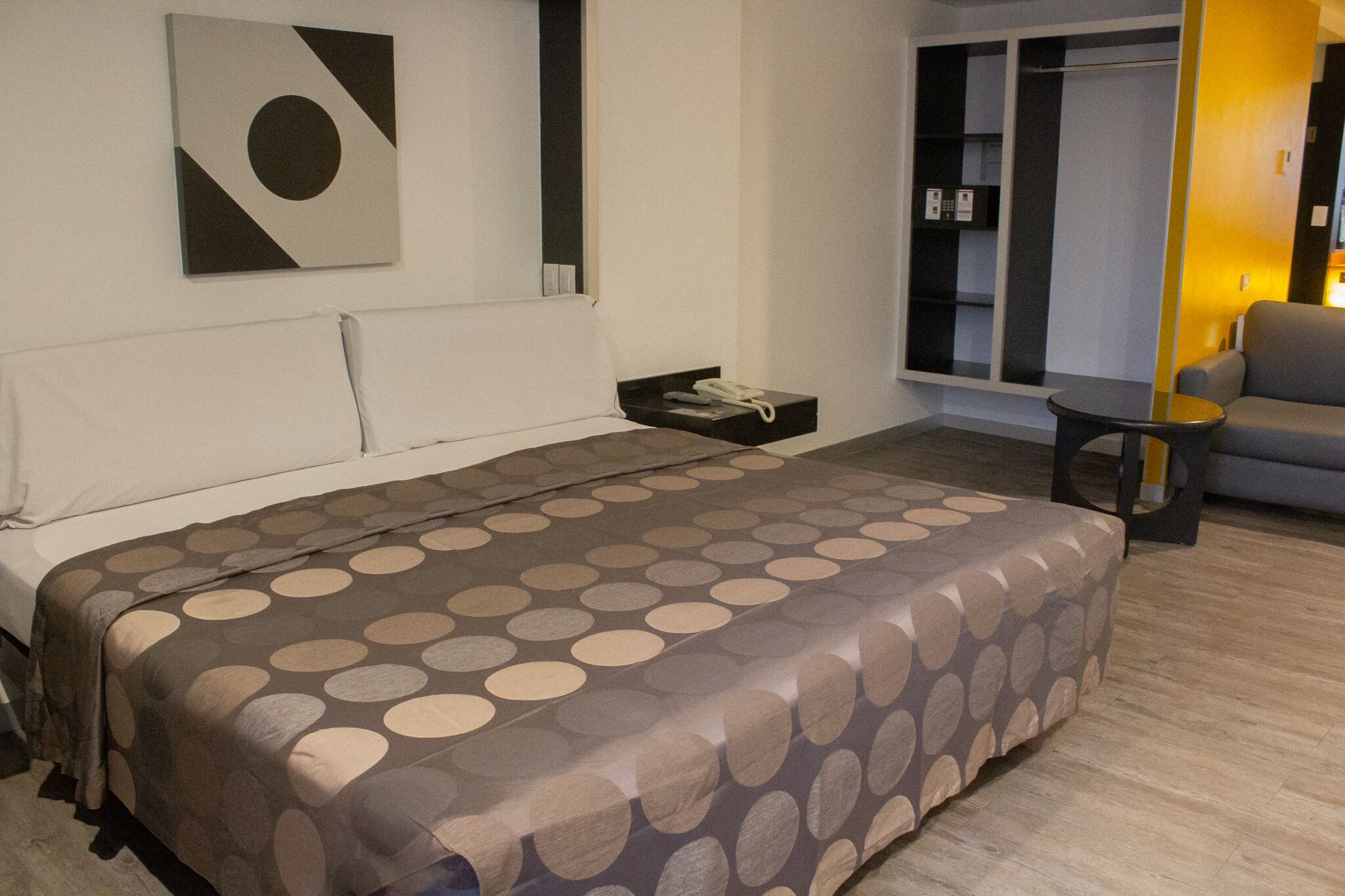 A neatly made bed with a patterned bedspread, a bedside table with a phone, and a chair in a modern room with a window and a wardrobe.