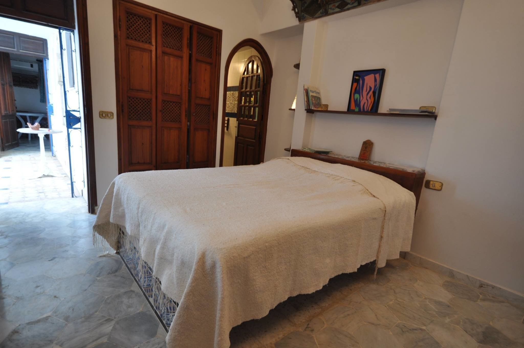 A bedroom with a bed, a wooden door, a small table, and a shelf with books.