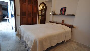 A bedroom with a bed, a wooden door, a small table, and a shelf with books.
