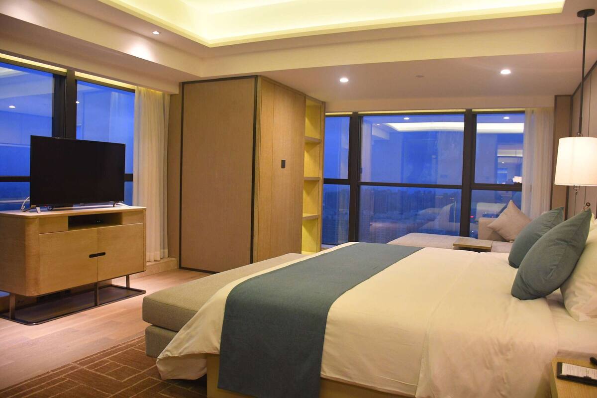 A modern hotel room with a large bed, a TV on a wooden cabinet, and a view of the city through large windows.