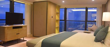 Quarto de hotel moderno com uma cama grande, uma televisão sobre um armário de madeira e vista da cidade através de janelas amplas.
