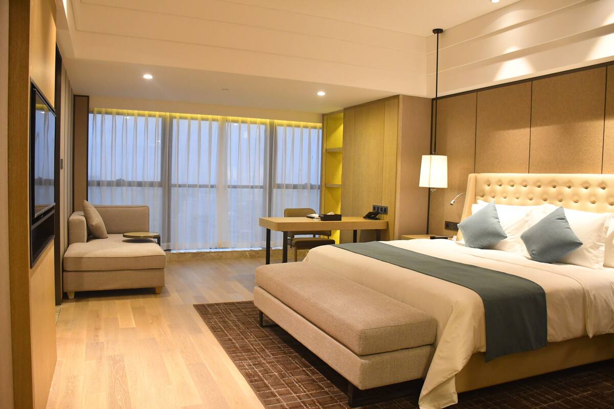 A hotel room with a large bed, a sofa, a desk, and a view of the city through large windows.