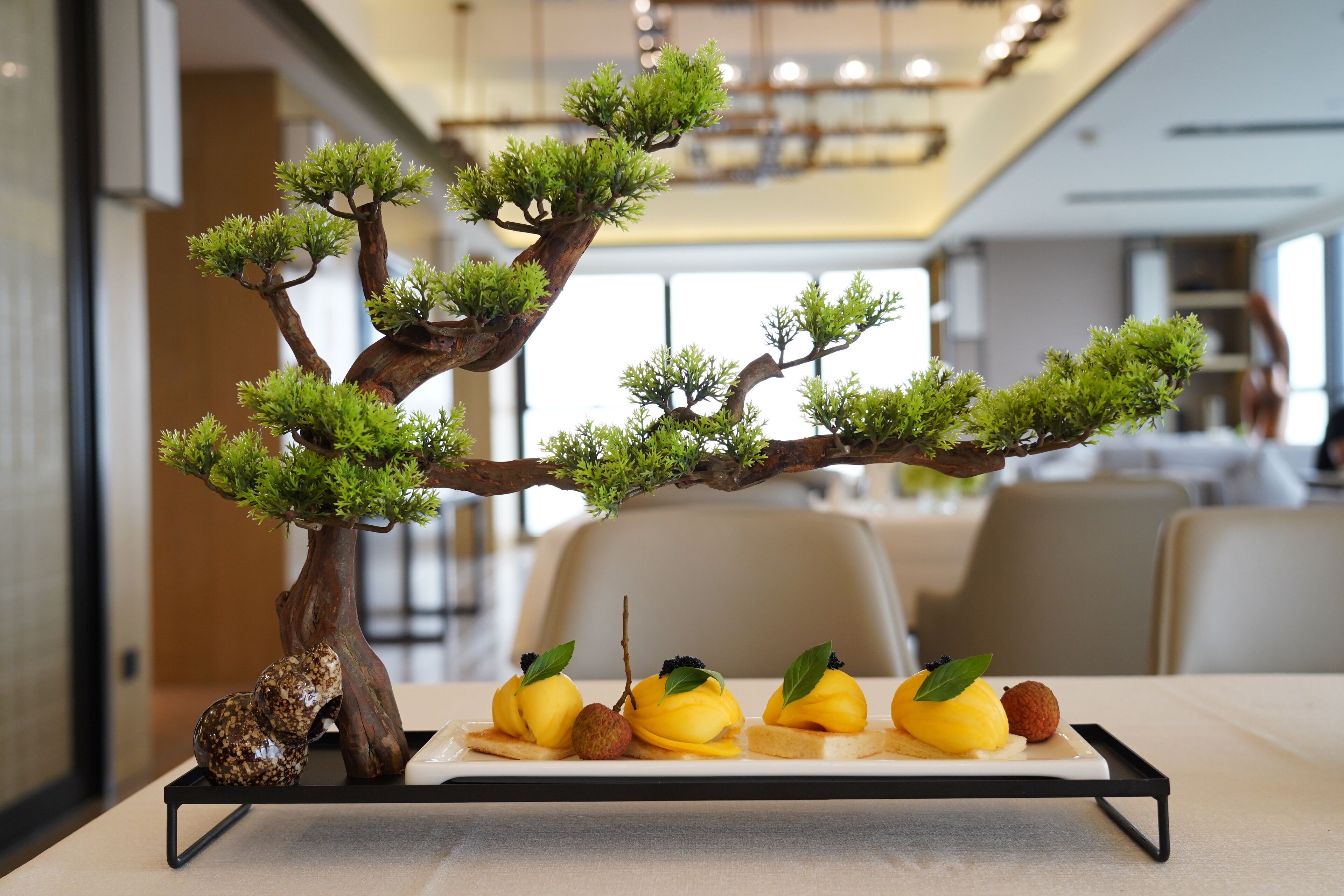 A bonsai tree with a tray of food items on a table in a modern interior setting.