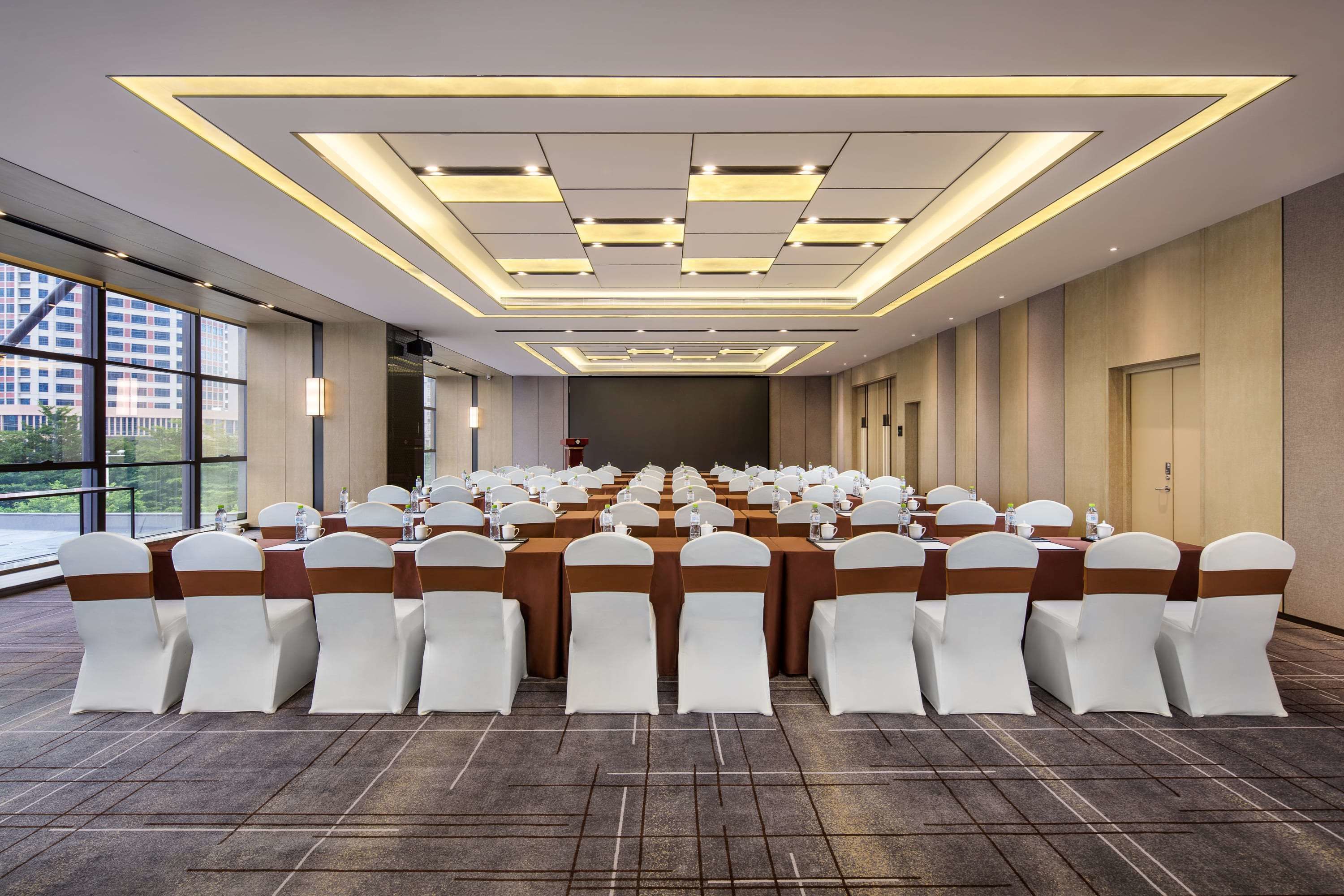 Une salle de conférence dotée d’une longue table, de chaises blanches ornées de rubans bruns et d’un plafond au design moderne.