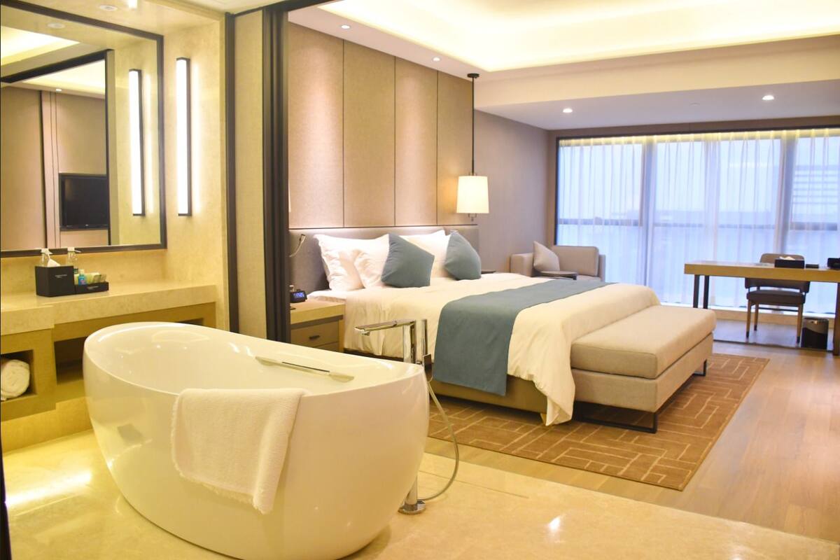 A modern hotel room with a large bathtub, a bed with multiple pillows, a desk, and a large window.