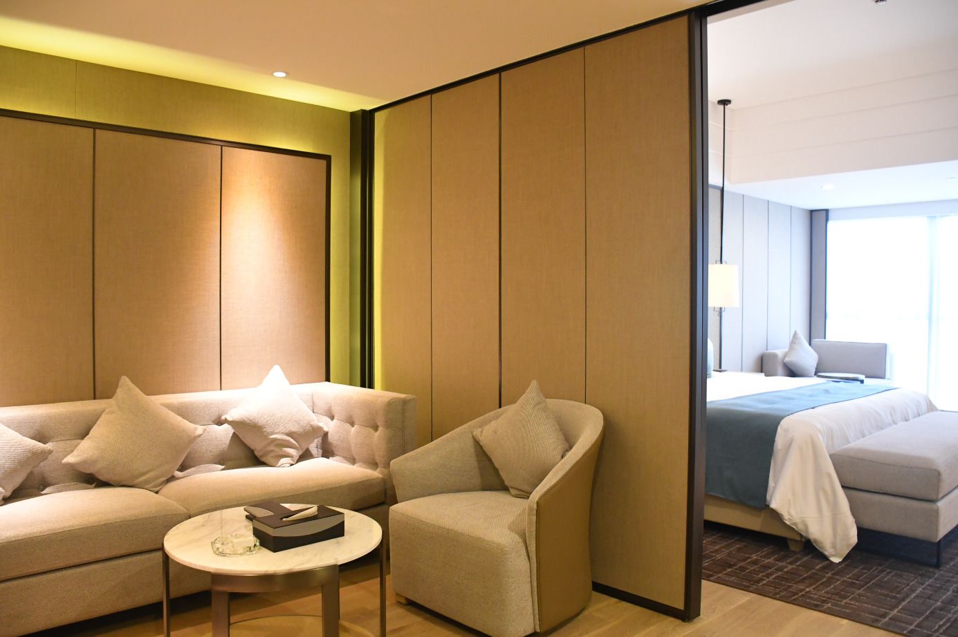 Een hotelkamer met een bank, fauteuil en een klein rond tafeltje. Er is een bed met een blauwe deken en een nachtkastje met een lamp.