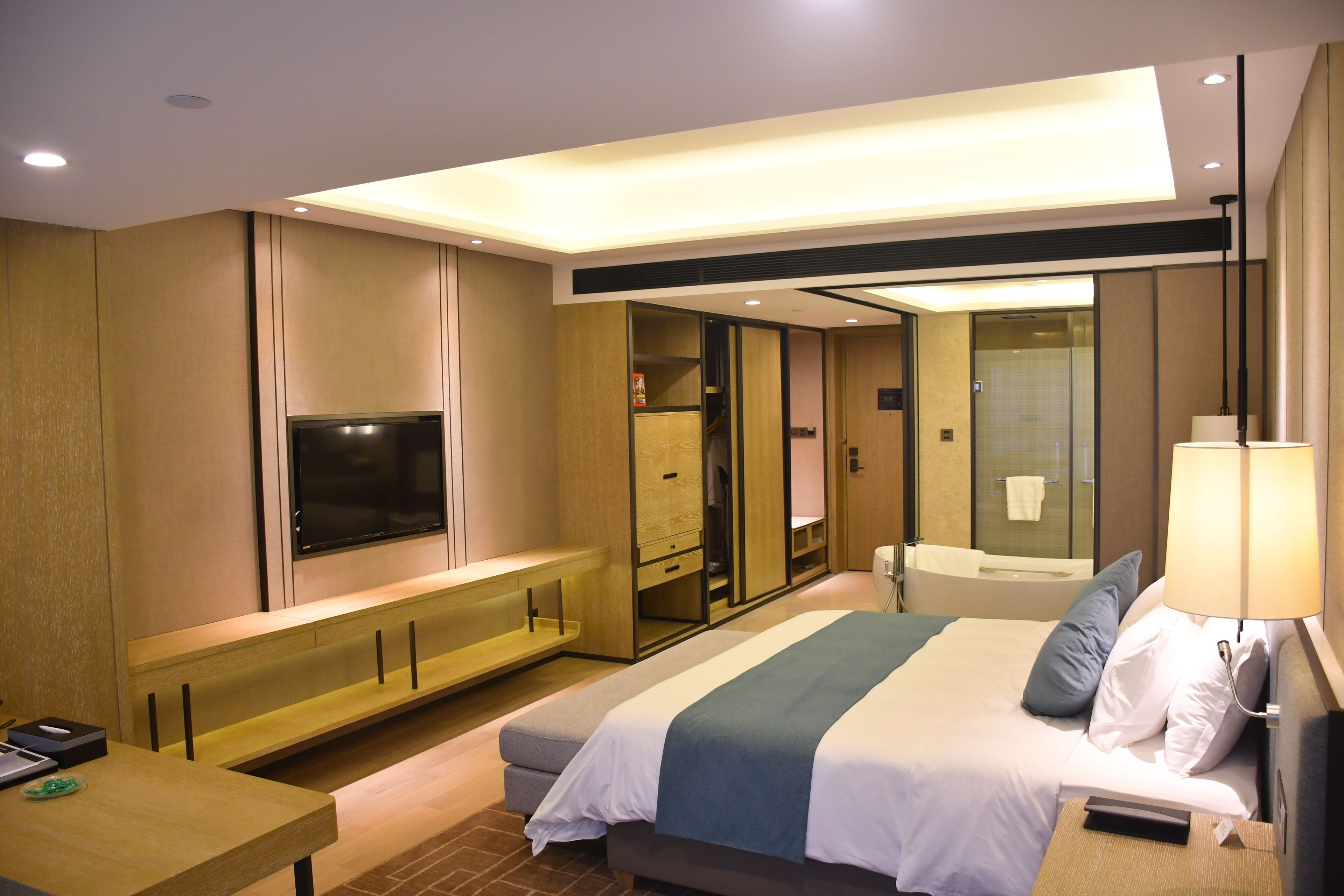 A hotel room with a bed, a TV, a bench, a desk, and a bathroom visible through an open door.