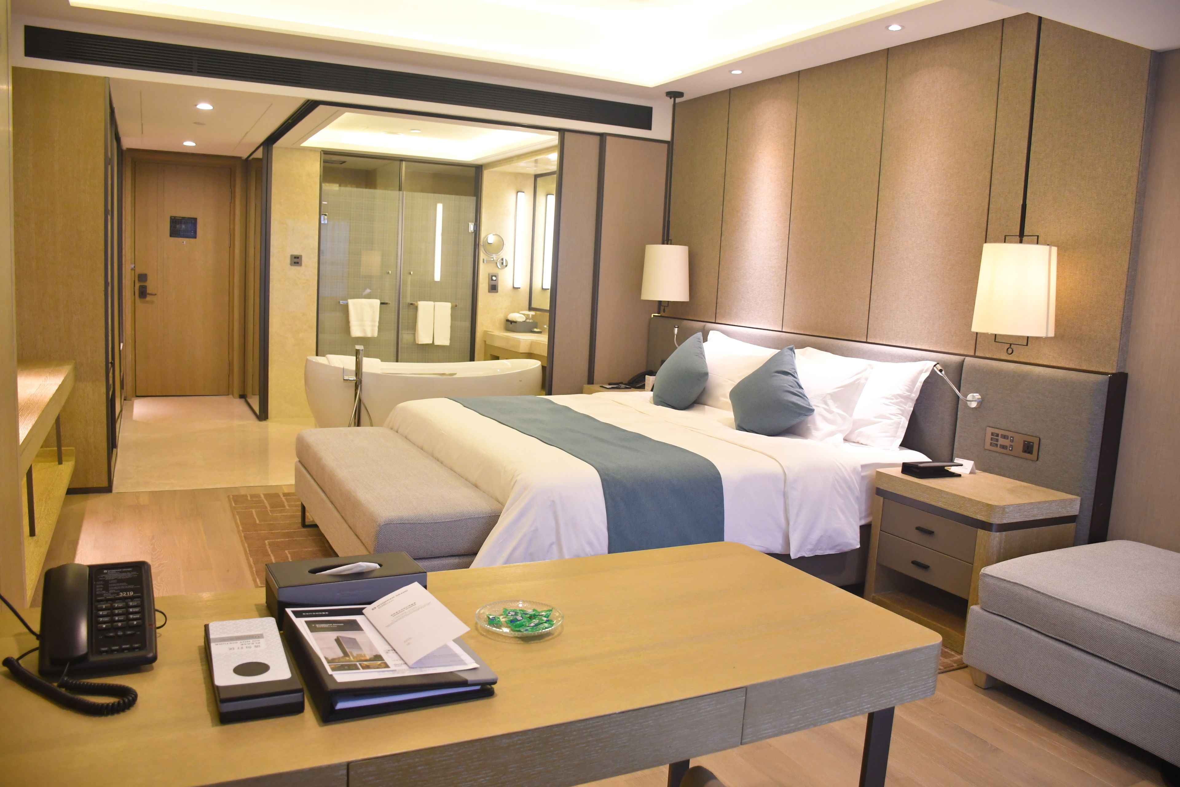 Ein Hotelzimmer mit Bett, Nachttisch, Schreibtisch und einem durch eine Glastür einsehbaren Badezimmer.