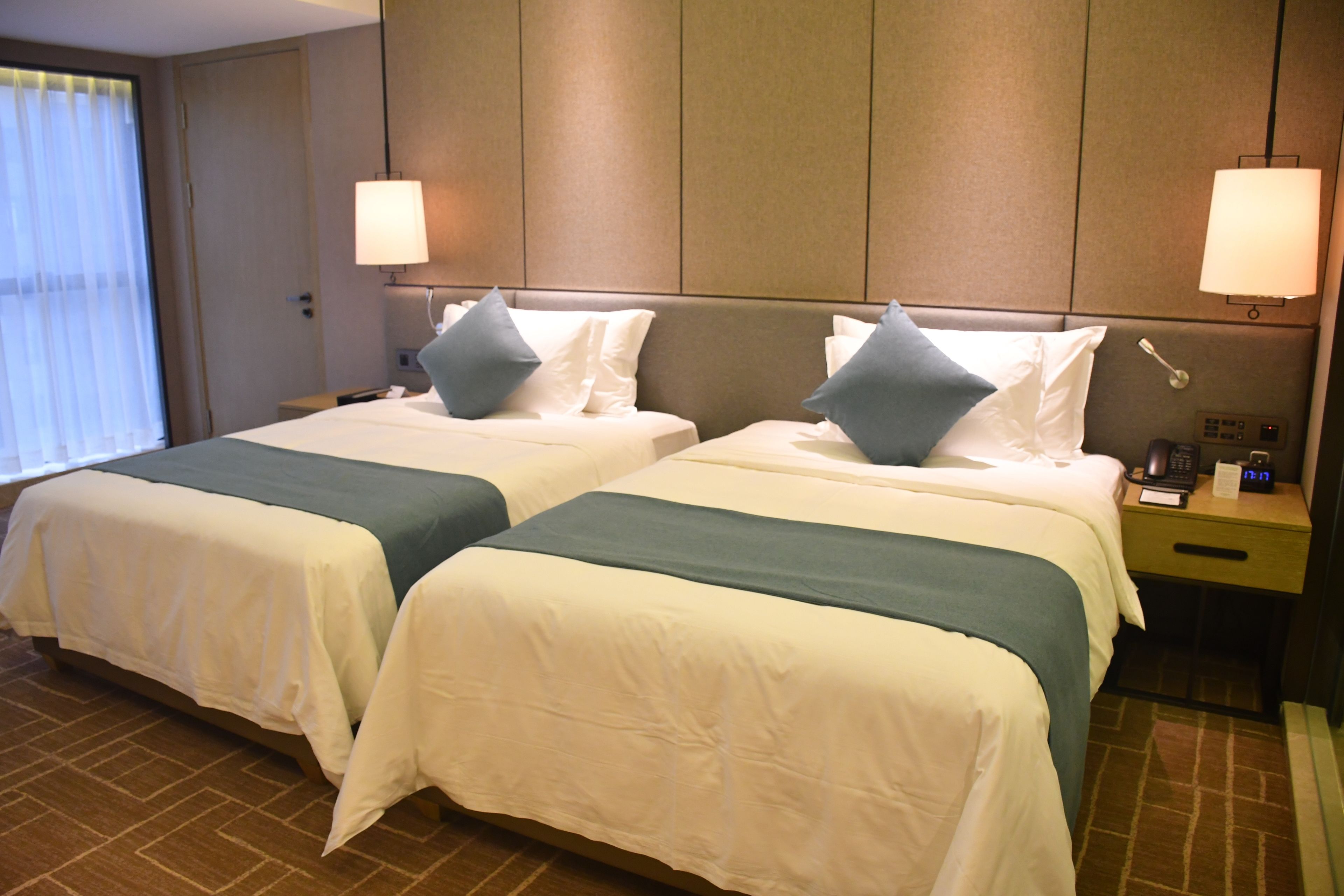A hotel room with two beds, each with white bedding and teal accents, a bedside table with a lamp, and a window with blinds.