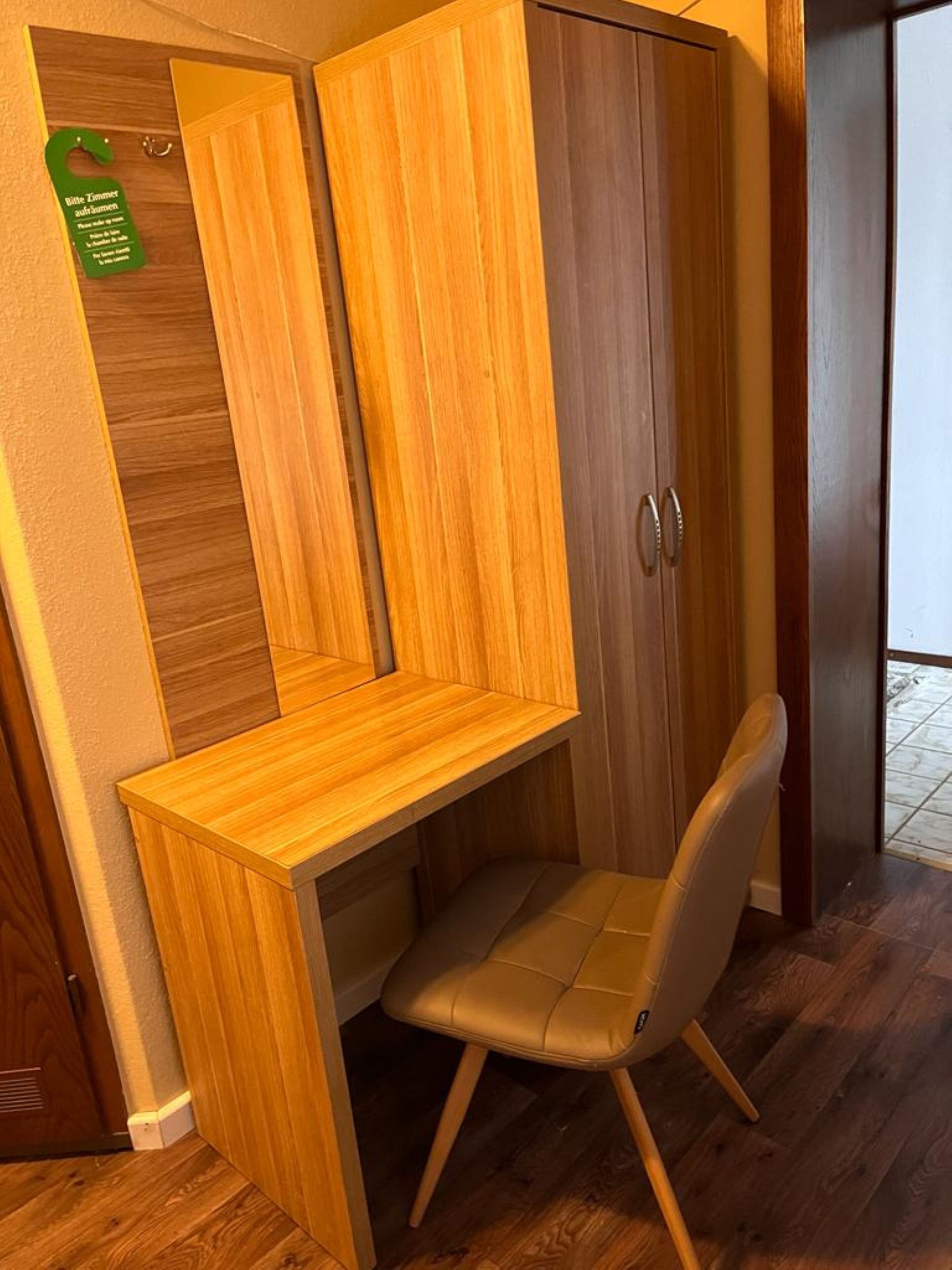 Une petite pièce comprenant un bureau en bois intégré, une chaise, une armoire avec un miroir et un panneau vert sur le mur.