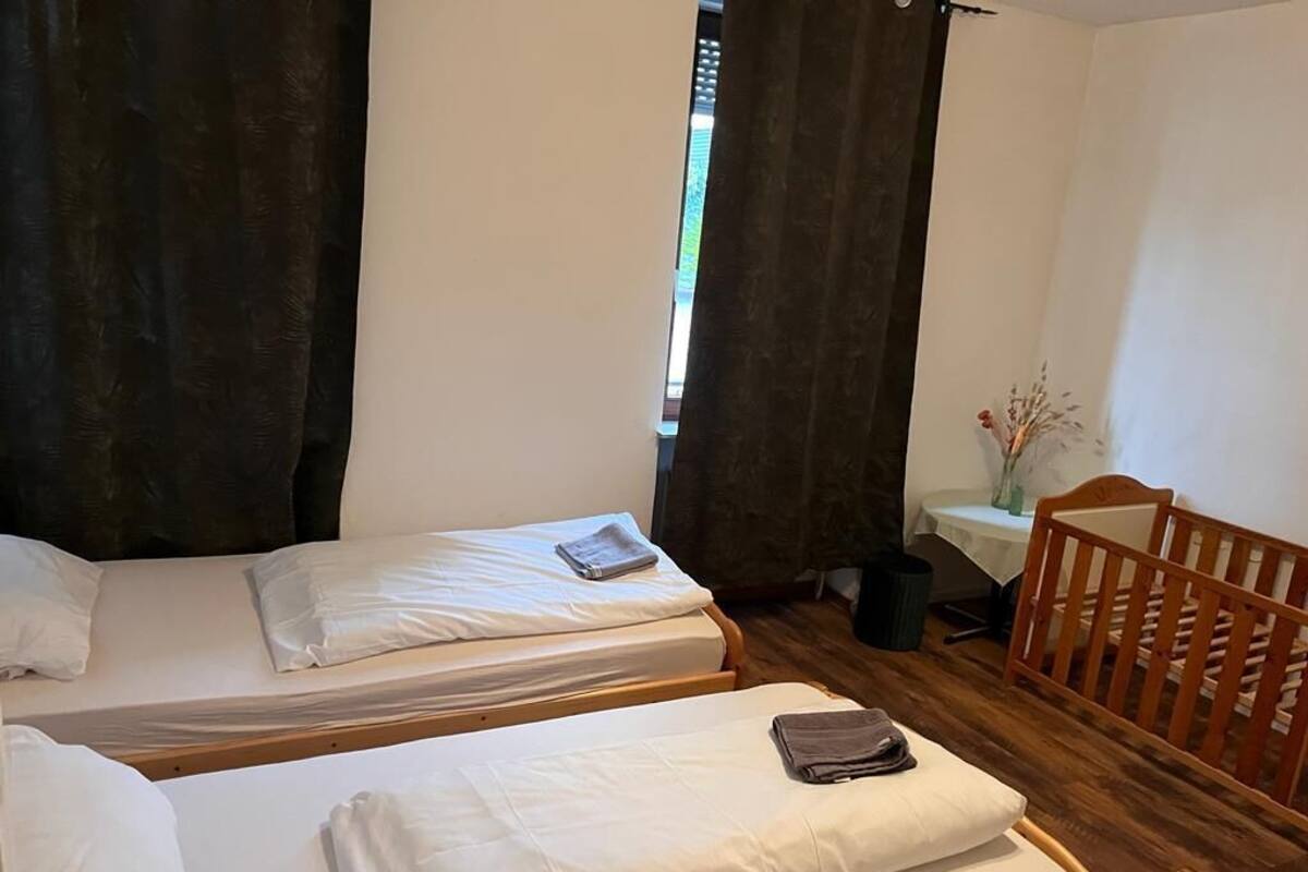 A room with two beds, a wooden crib, a small table with a vase, and a window with curtains.