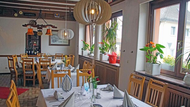 A dining area with wooden chairs and tables set for a meal, featuring a large window with plants and a view of buildings outside. - Hotel zur Post (Hünfelden)