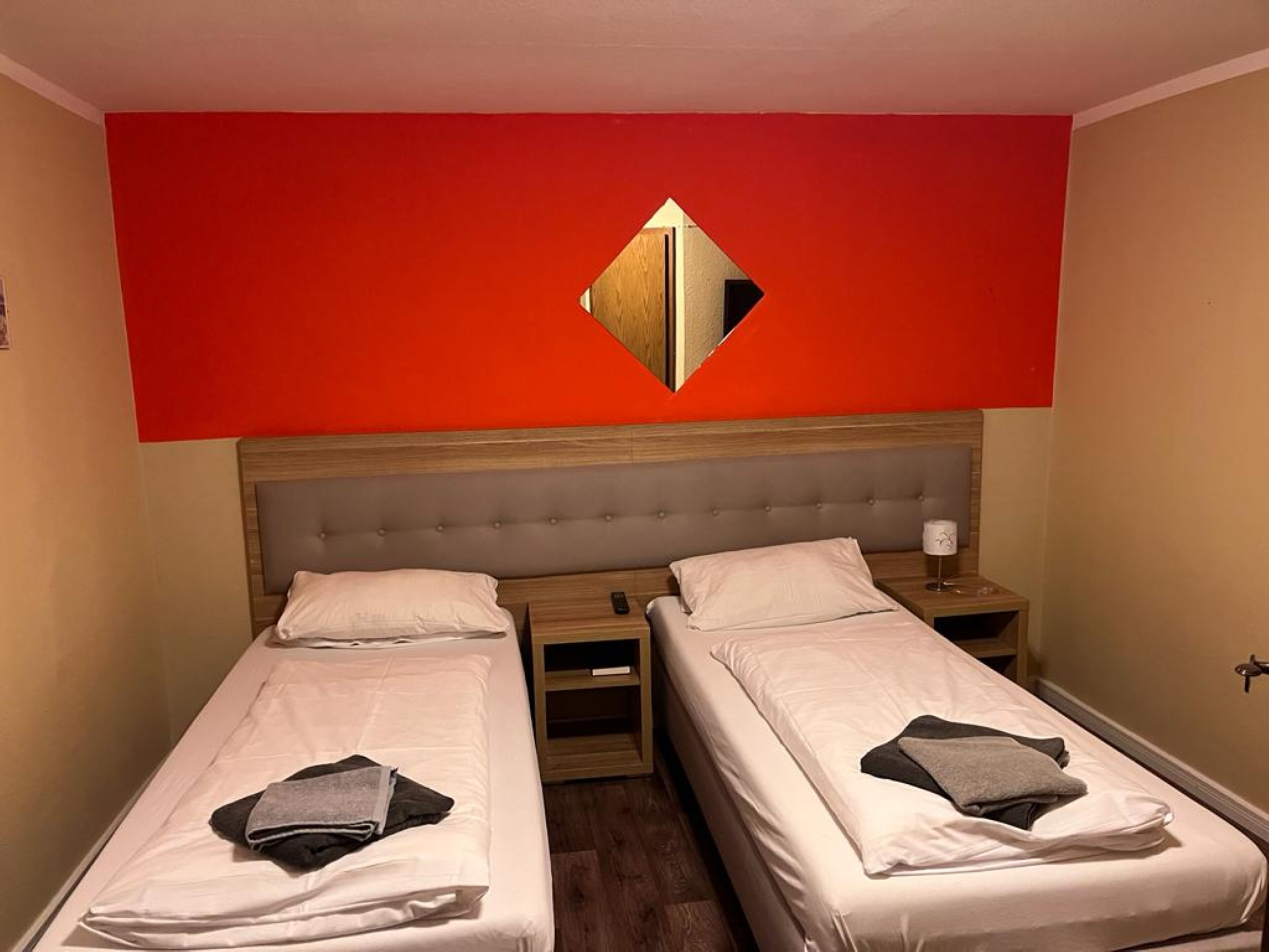 A hotel room with two single beds, a wooden headboard, a red accent wall, and a mirror.