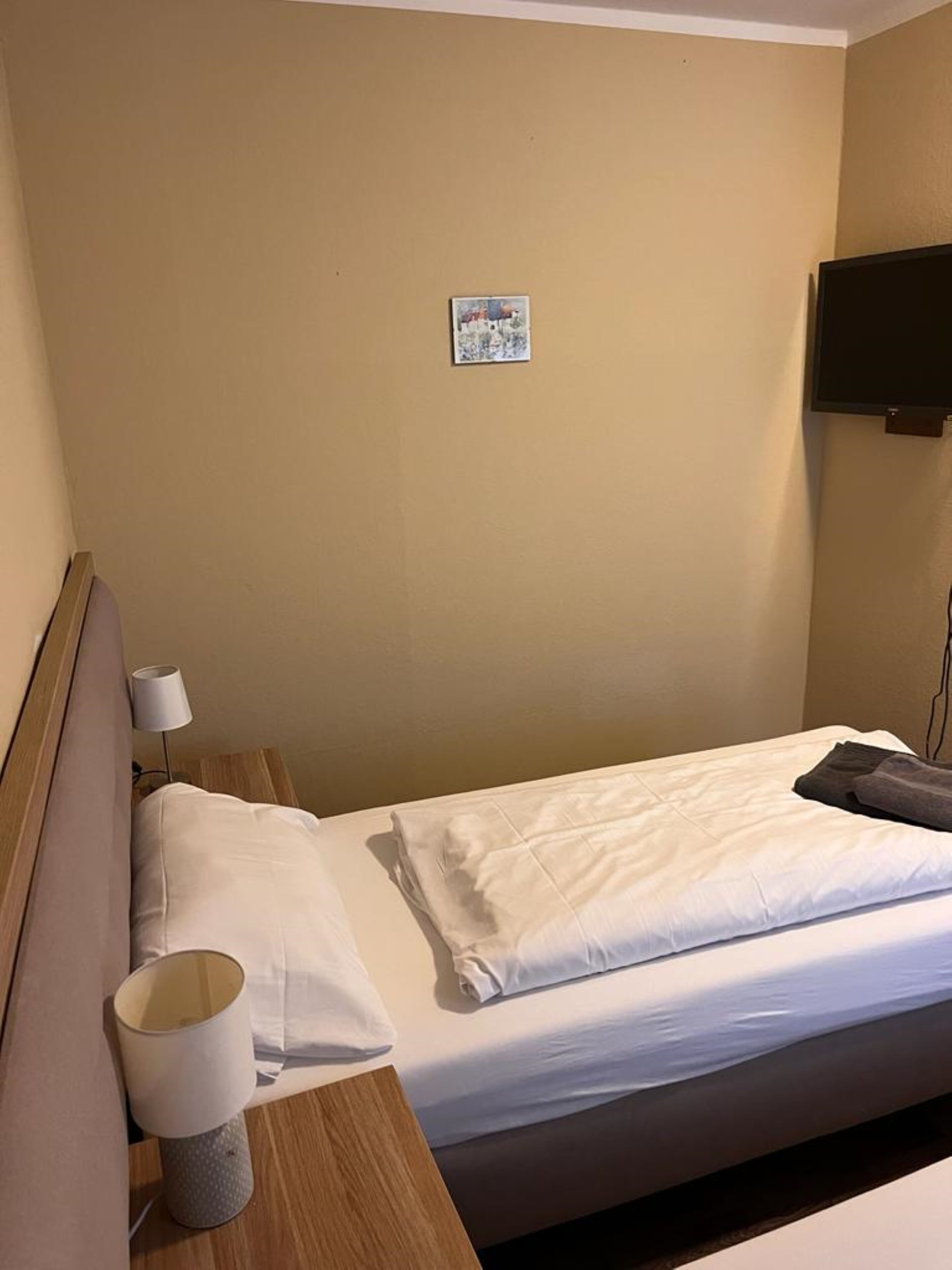 Ein Hotelzimmer mit einem Einzelbett, einem Nachttisch mit Lampe und einem an der Wand befestigten Fernseher.