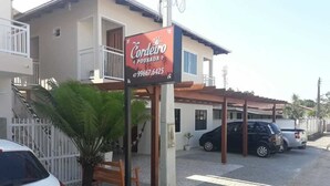 A building with a sign for 'Cordeiro Pousada' and a contact number. There are parked cars and a palm tree in front. - Pousada Cordeiro (Penha)