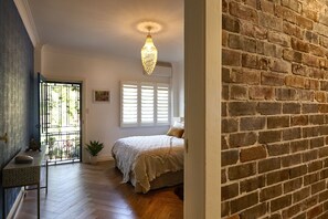 Un dormitorio con cama, mesita de noche, planta en maceta y ventana con contraventanas.