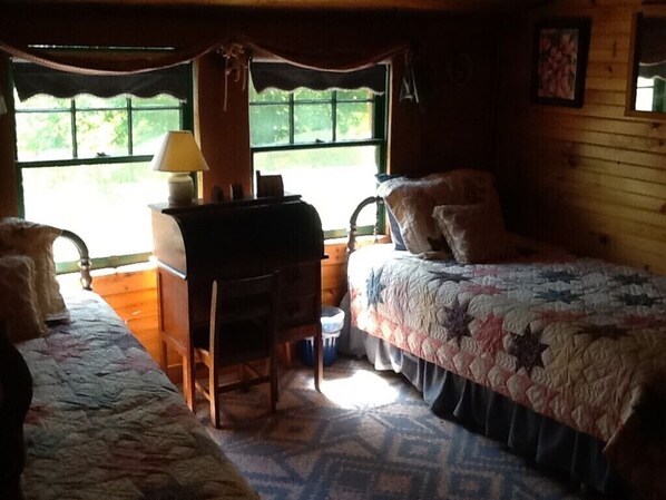 A cozy cabin room with two beds, a desk, and a window providing a view of greenery outside.