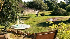 A round table with a bottle and glasses, surrounded by greenery and a view of a grassy area with trees.