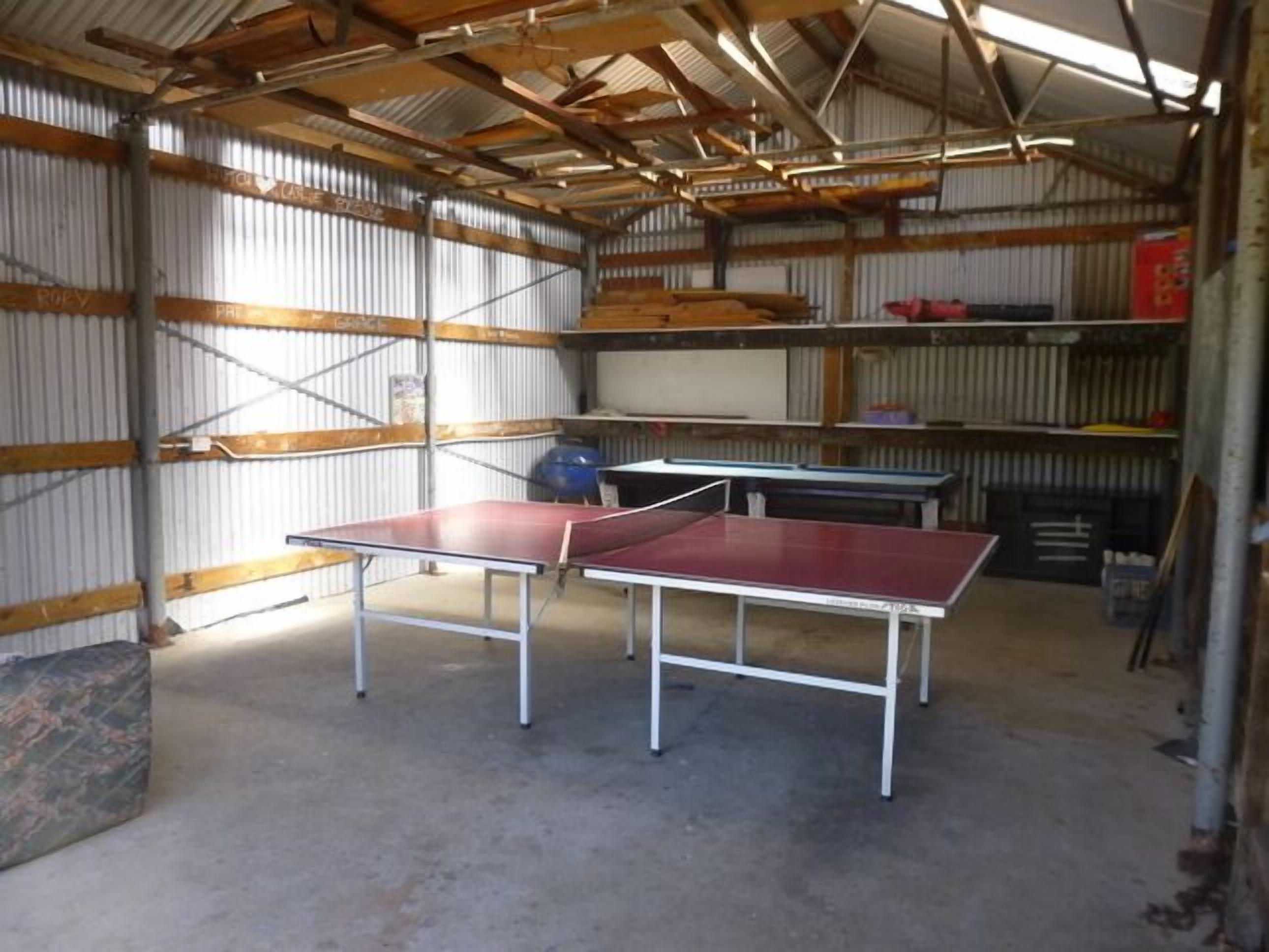 A spacious indoor area with two ping pong tables, metal shelving, and a corrugated metal roof.