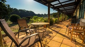 An outdoor patio with a round table and chairs, surrounded by stone tiles and greenery, under a wooden pergola.