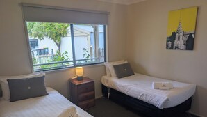 A hotel room with two beds, a nightstand with a lamp, a window with a view of greenery, and a painting on the wall. - Riverside Golf Club 3 Bedroom Holiday House (Nichols Point)