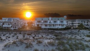 Un hotel frente a la playa con edificios de varios pisos, una playa arenosa en primer plano y el sol poniéndose al fondo.