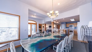 A spacious dining area with a large table and multiple chairs, a chandelier, and a kitchen in the background. - Cloud Nine - KDH MP 7, Semi-Oceanfront, Ocean View, 120 Yds to Beach, Pool, Hot Tub, Pets OK (Kill Devil Hills)