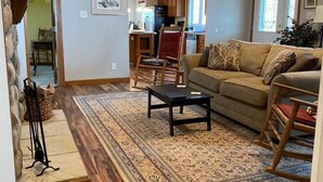 A spacious living room with a large patterned rug, wooden flooring, a sofa, and a rocking chair. A kitchen is visible in the background. - Escape To The Country In Comfort And Style In An Eco-Friendly House (Rushville)