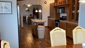 A dining area with a round wooden table and four chairs, a kitchen with wooden cabinets and a microwave, and a hallway leading to a living room with a sofa and a coffee table. - Escape To The Country In Comfort And Style In An Eco-Friendly House (Rushville)