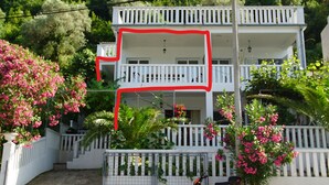 A multi-story building with balconies, surrounded by lush greenery and pink flowering plants.