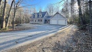 Une maison à deux étages avec un garage, entourée d’une allée en gravier et d’une zone boisée.