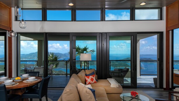A modern living room with a large sofa, dining table, and chairs, large windows offering ocean views. - The Crown Jewel of Calichi (St. John)