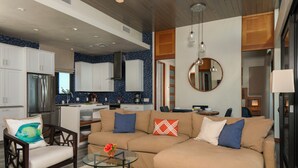 A modern living room with a beige sectional sofa, a glass coffee table, and a dining area with a kitchen in the background. - The Crown Jewel of Calichi (St. John)