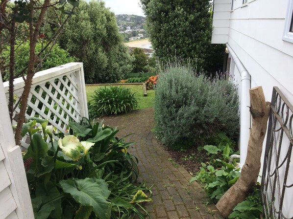 A paved garden path leading to a white fence, surrounded by lush greenery and a view of a distant town. - Gorgeous beach house on Oneroa Bay (Little Oneroa)