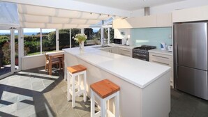 Cocina moderna con una isla grande, encimeras blancas y electrodomésticos de acero inoxidable. Hay dos taburetes de madera y una pequeña mesa con sillas.