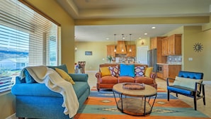 A spacious living room with a blue sofa, a coffee table, and a view through large windows. - La Verkin Home w/ Hot Tub, Kayaks, & Bikes! (La Verkin)