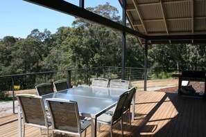 An outdoor dining area with a glass table and metal chairs, surrounded by a wooden deck and a metal railing, overlooking a forested area. - Yallingup Hidden Valley (Yallingup)