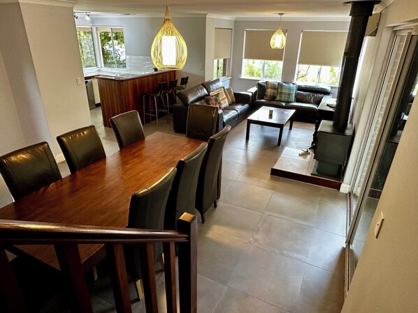 A spacious living area with a wooden dining table, leather chairs, a sofa, and a coffee table. There are two hanging lights and a kitchen area in the background.