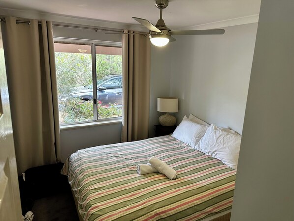 A hotel room with a bed, a ceiling fan, a window with curtains, a nightstand with a lamp, and a view of a parked car outside.