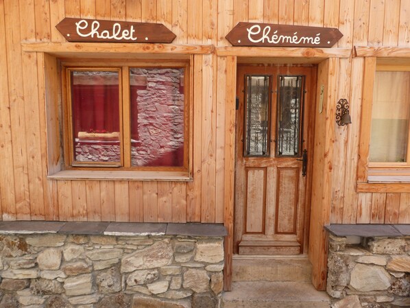 Un bâtiment en bois avec des fondations en pierre, une porte en bois dotée de panneaux de verre et deux panneaux au-dessus de la porte indiquant les mots « Chalet » et « Chéméné ».