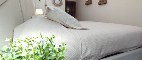 Una cama bien hecha con edredón y almohadas blancos, una planta en maceta al lado, y una pared con un espejo circular y dos pequeñas repisas con objetos decorativos.