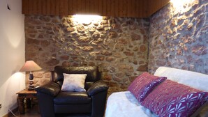 Una habitación acogedora con pared de piedra, un sillón de cuero con cojín, un sofá blanco con cojines estampados y una mesita con lámpara.