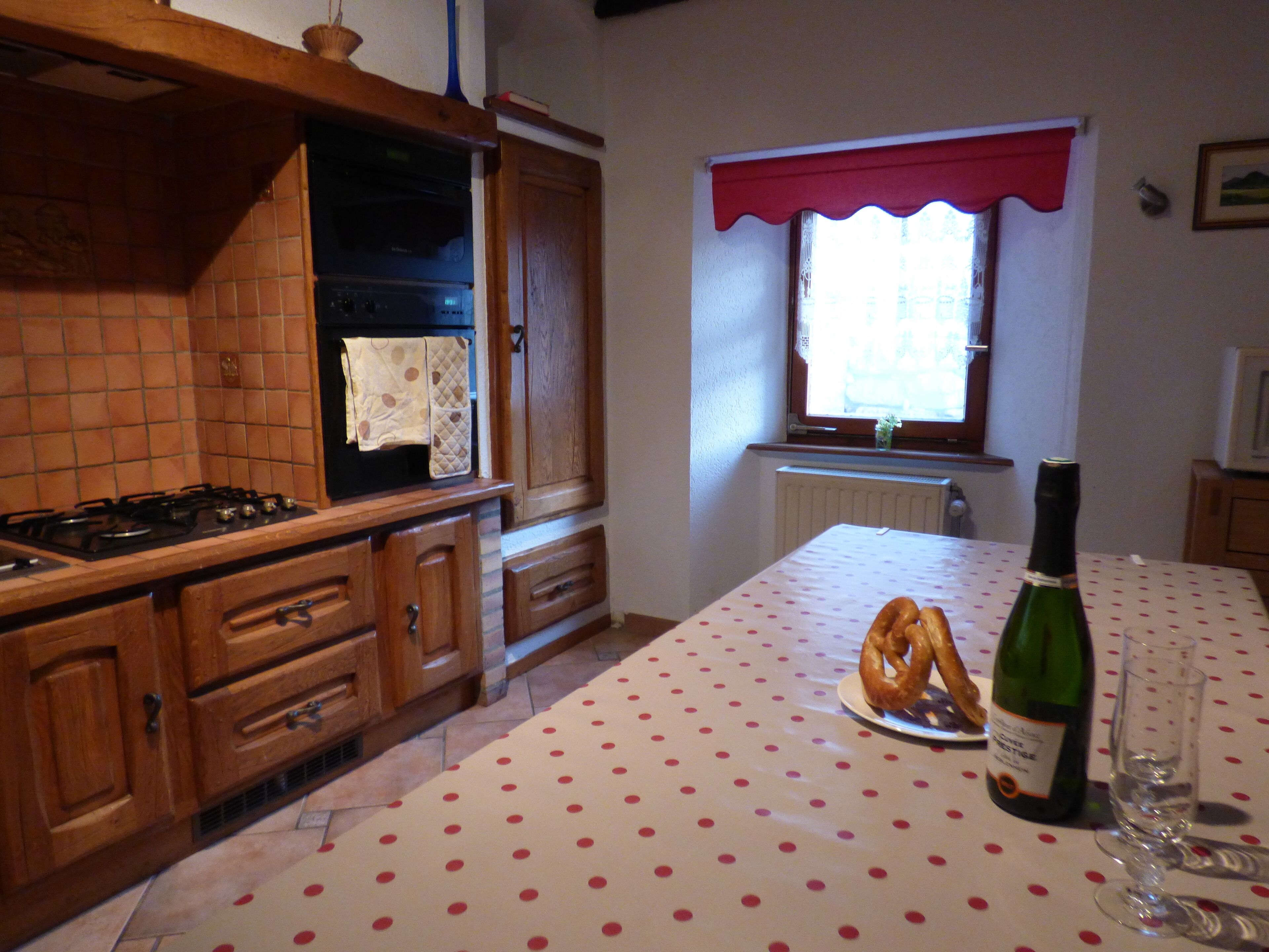 Une cuisine avec des meubles de rangement en bois, une cuisinière, une fenêtre agrémentée d’une lambrequin rouge et une table recouverte d’une nappe à pois.