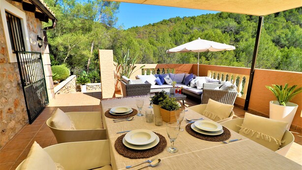 An outdoor dining area with a table set for two, surrounded by wicker furniture and a view of a forested hillside.
