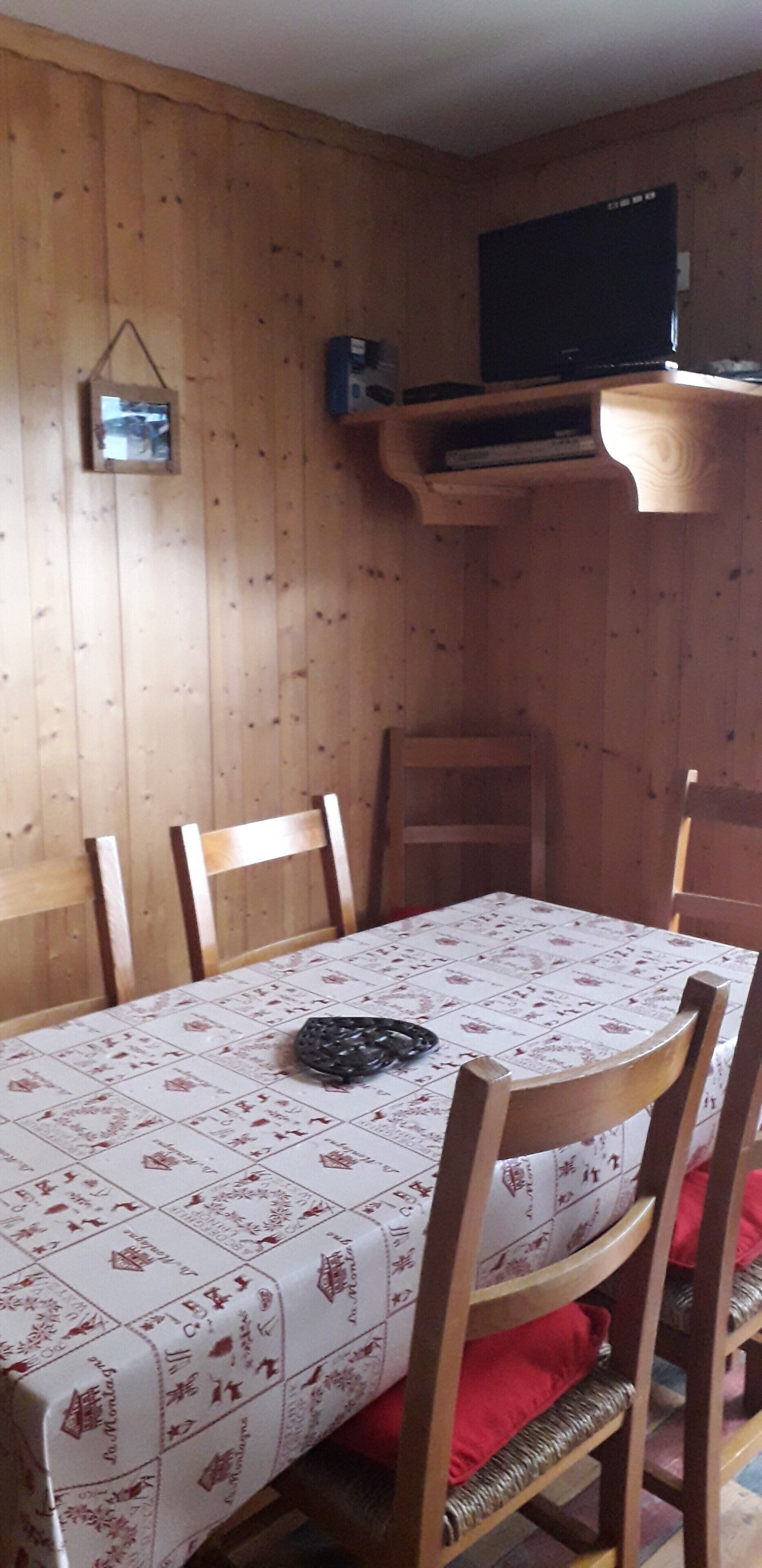 Een eethoek met een tafel bedekt met een tafelkleed met patroon, houten stoelen en een televisie bevestigd aan een houten plank.