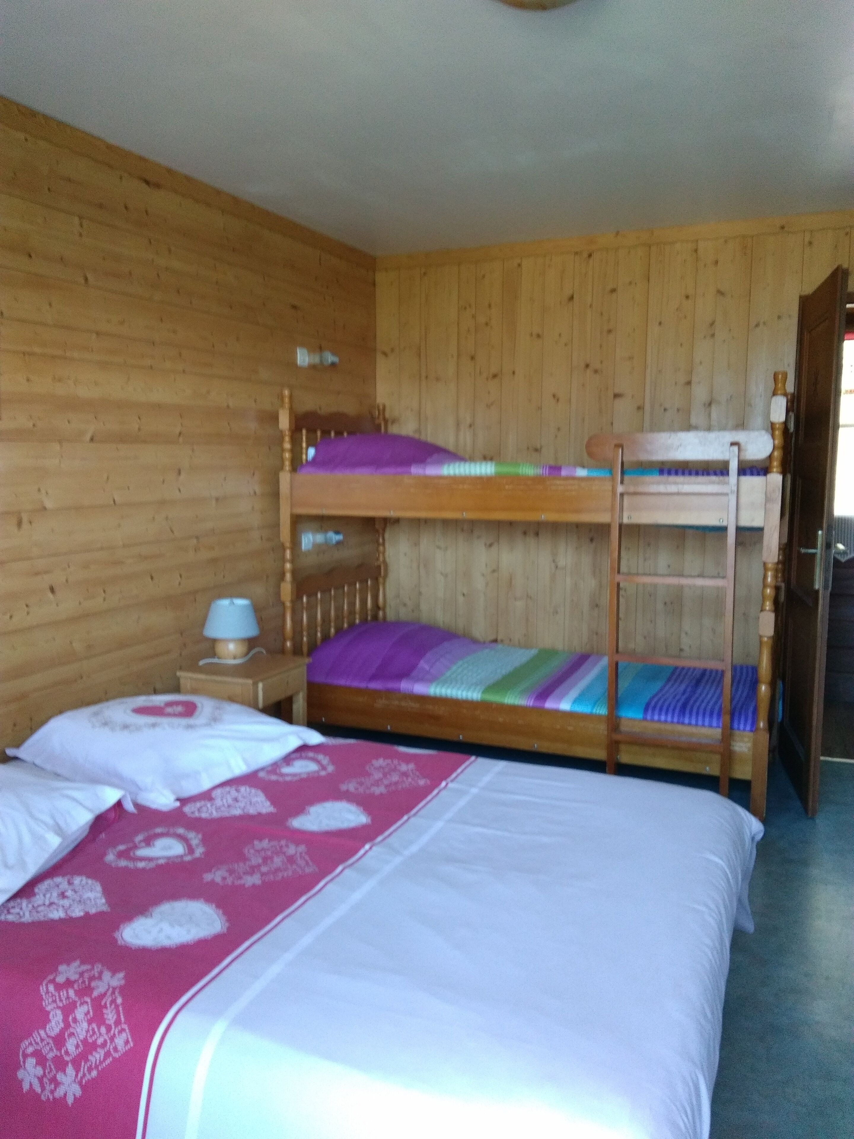 A wooden room with bunk beds, a bedside table with a lamp, and a bed with a patterned blanket.