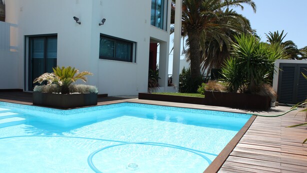 Ein Schwimmbad mit Holzterrasse, umgeben von Topfpflanzen und einem modernen Gebäude mit großen Fenstern.